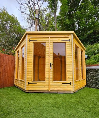 A small wooden garden shed with large windows, set in a landscaped area.